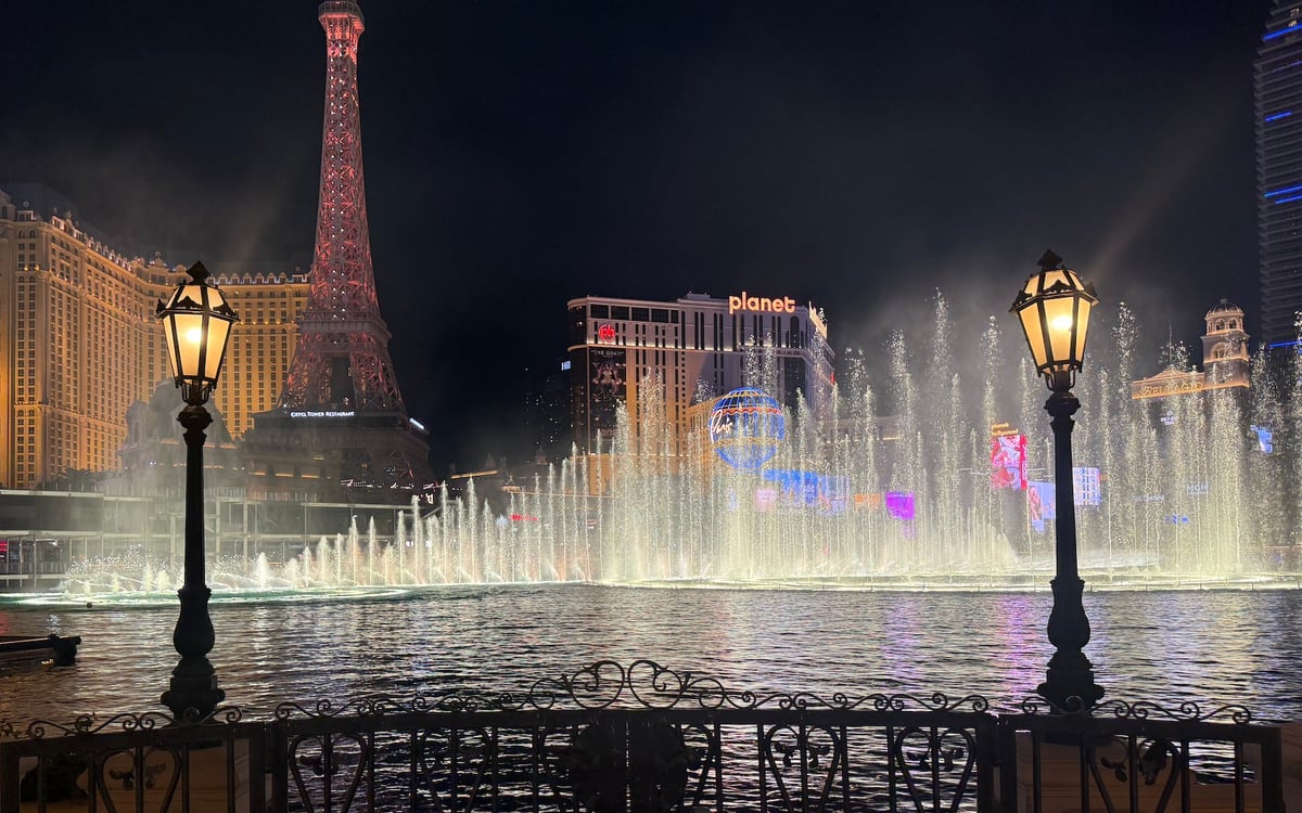 Night view of the Bellagio Fountains in Las Vegas with colorful illuminated water jets, the replica Eiffel Tower, and vintage street lamps in the foreground